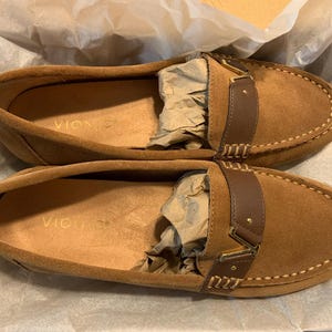 May include: A pair of brown suede loafers with a leather strap and gold-colored buckle. The shoes have a stitched design around the edges and are displayed on white tissue paper. The brand name "Vionic" is visible inside the shoe.