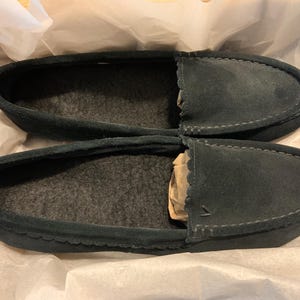 May include: A pair of dark teal suede moccasin-style slippers. The slippers have a soft, gray interior and are packaged in white tissue paper. The stitching is visible around the edges of the toe and heel.