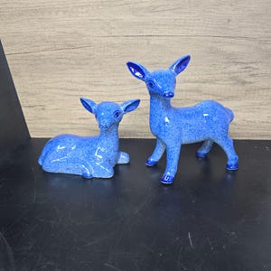 May include: Two ceramic deer figurines with a speckled blue glaze. One deer is in a reclining position with its legs tucked underneath, while the other deer is standing on all four legs. Both figures feature pointed ears and dark blue eyes.