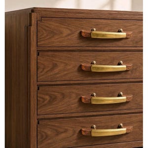 May include: A wooden chest of drawers featuring four horizontal drawers with a visible wood grain pattern. Each drawer is equipped with a handle consisting of a central brushed brass metal plate flanked by two dark wood end caps.