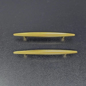 May include: Two elongated, gold-toned metal cabinet pulls featuring a smooth, tapered design with a central ridge. Each handle is supported by two small, cylindrical posts that extend outward, providing a sleek and uniform appearance for furniture.