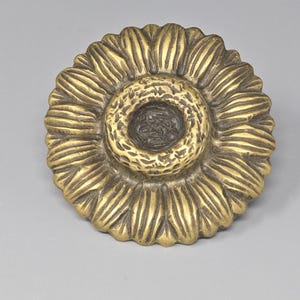 May include: A circular brass decorative object shaped like a sunflower with a textured, raised center and radiating petals. The metal has a polished gold-tone finish with darkened recessed areas between the petals to highlight the detailed design.