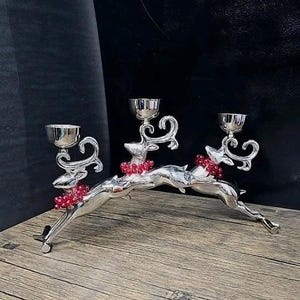 May include: A silver-toned metal candelabra featuring three leaping reindeer figures. Each reindeer has a small candle cup mounted on its back and wears a decorative collar made of small, shiny red beads. The structure is arched and metallic.