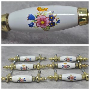 May include: A set of six drawer pulls featuring white ceramic handles with a floral pattern of pink, blue, and yellow flowers. Each handle is accented with gold-toned metal hardware at both ends, including mounting screws for installation.