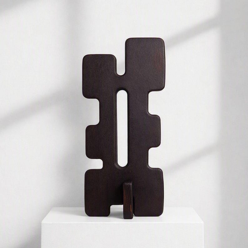 Tall black sculpture with abstract shape and black distressed textured finish. The modern sculpture is standing on top of a white pedestal against a white background.