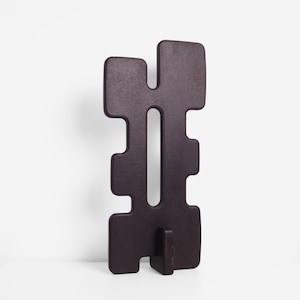 Image of the TREZE sculpture in matte black, shown slightly turned to the side to reveal its geometric contours and the elongated central cutout. The sculpture stands upright on its matching matte black base, placed on a clean white surface against a plain white background.