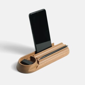 A black smartphone docked in a light brown wooden desk organizer with 3 pen slots and a small hollow area for paperclips.