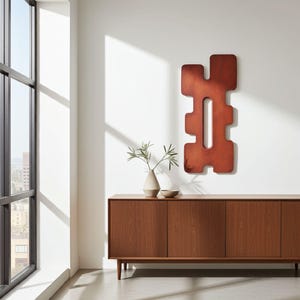 Reddish-brown abstract wall sculpture with geometric cutouts, vertically mounted above a mid-century modern wooden sideboard. Handcrafted contemporary art piece in a minimalist interior with ceramic vases, greenery, and city view. Ideal for modern apartments, curated design spaces, or art-focused interiors.