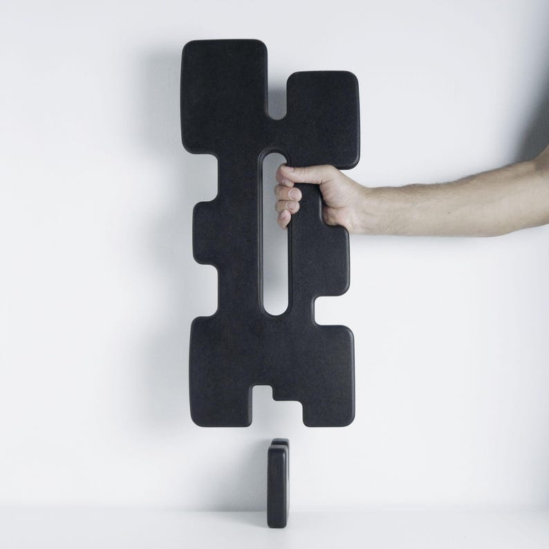 Image of the TREZE sculpture in matte black, with a hand holding the tall geometric piece featuring a central elongated cutout. Below it, the matching matte black base — the support that allows the sculpture to stand upright — rests on a white surface against a clean white background.