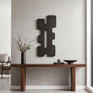 Modern minimalist interior featuring a large black abstract wall sculpture with a geometric symmetrical design and central vertical opening, displayed above a wooden console table styled with books, a dark vase with branches, and decorative objects. Contemporary wall art for sophisticated home decor.