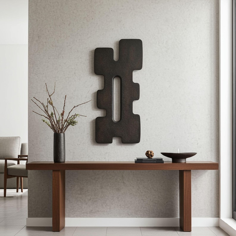 Modern minimalist interior featuring a large black abstract wall sculpture with a geometric symmetrical design and central vertical opening, displayed above a wooden console table styled with books, a dark vase with branches, and decorative objects. Contemporary wall art for sophisticated home decor.