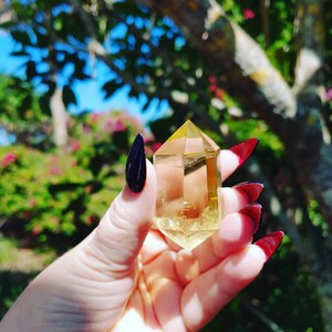May include: A clear, yellow citrine crystal point with a faceted shape. The crystal is being held in a hand with black nail polish.