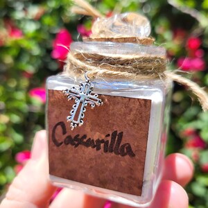 May include: A small glass jar with a cork stopper and a silver cross charm. The jar is tied with twine and has a label that reads "Cascarilla".