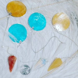 May include: A set of five dowsing pendulums with chains. The pendulums are made of different materials, including metal, crystal, and wood. The pendulums are in various colors, including yellow, blue, and white.