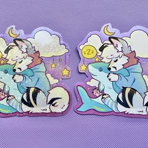 May include: Two identical die-cut stickers featuring a white and grey anthropomorphic canine character with a blue hoodie, sleeping while hugging a light blue shark plush. The background includes yellow stars, clouds, a crescent moon, and Zzz text.
