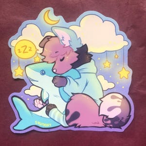 May include: A die-cut sticker featuring a stylized illustration of a fluffy, pink-furred creature wearing a light blue hoodie, sleeping while hugging a large, light blue shark plush. The background includes yellow stars, clouds, and a crescent moon.