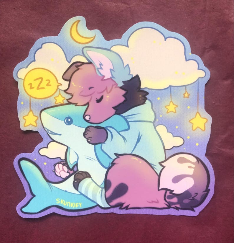 May include: A die-cut sticker featuring a stylized illustration of a fluffy, pink-furred creature wearing a light blue hoodie, sleeping while hugging a large, light blue shark plush. The background includes yellow stars, clouds, and a crescent moon.