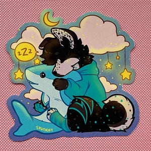May include: A die-cut sticker featuring a black and white anthropomorphic creature with a teal hoodie hugging a light blue shark. The background shows yellow stars, clouds, a crescent moon, and a yellow circle with the text ZZZ. The text SKUNKIFY is visible.