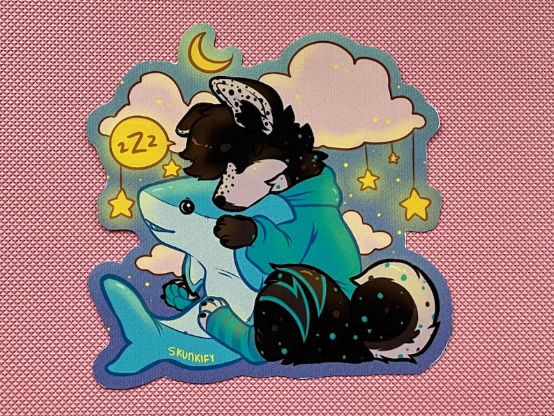 May include: A die-cut sticker featuring a black and white anthropomorphic creature with a teal hoodie hugging a light blue shark. The background shows yellow stars, clouds, a crescent moon, and a yellow circle with the text ZZZ. The text SKUNKIFY is visible.