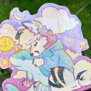 May include: A holographic sticker featuring a stylized anthropomorphic animal with light fur wearing a blue hoodie, sleeping while hugging a blue shark plush toy. The background includes a crescent moon, clouds, and stars with a ZZZ speech bubble.