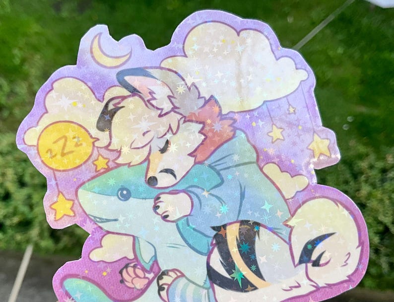 May include: A holographic sticker featuring a stylized anthropomorphic animal with light fur wearing a blue hoodie, sleeping while hugging a blue shark plush toy. The background includes a crescent moon, clouds, and stars with a ZZZ speech bubble.