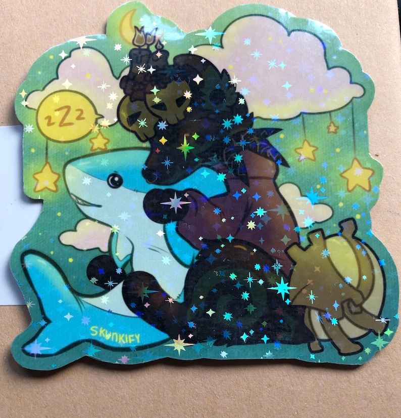 May include: A holographic sticker featuring a dark, shadowy creature with skull-like features embracing a light blue shark. The background includes yellow stars, clouds, a crescent moon, and a yellow circle with the text zzz. The artist name Skunkify is visible.