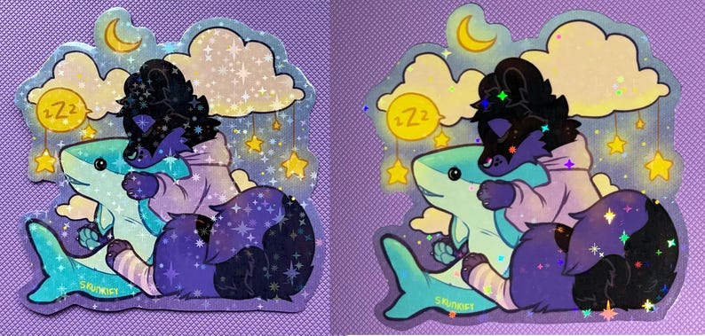 May include: A holographic sticker featuring a dark purple and black creature wearing a light purple hoodie, hugging a light blue shark. The background includes yellow clouds, a crescent moon, hanging stars, and a yellow circle with Zz text.