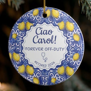 May include: A round ceramic ornament featuring a blue and white Mediterranean-style tile pattern with yellow lemons. The center text reads Ciao Carol! FOREVER OFF-DUTY, with a small stethoscope icon and the text RETIRED 2026, hanging by a gold cord.