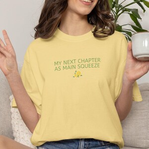 Op de afbeelding: Lichtgele t-shirt met de groene tekst "MY NEXT CHAPTER AS MAIN SQUEEZE" en een citroenafbeelding. Het shirt wordt gecombineerd met een spijkerbroekshort. De persoon houdt een witte mok vast.