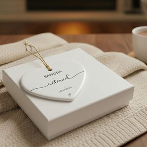 May include: A white heart-shaped ceramic ornament featuring the name SANDRA, the word retired in script, the date 09.15.2026, and a small heart icon. The ornament includes a gold hanging string and rests on a white square gift box.