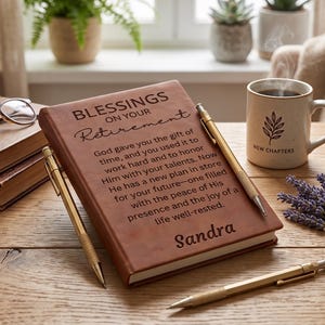 May include: A brown leather-bound journal with the text Blessings on your retirement and the name Sandra engraved on the cover. The journal is accompanied by two gold-toned pens, a white ceramic mug with a leaf graphic, and a pair of eyeglasses.