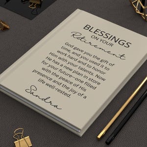 May include: A beige journal with the words "Blessings on Your Retirement" and a personalized name. The journal is accompanied by gold binder clips, pencils, and decorative geometric shapes, all set on a dark surface.
