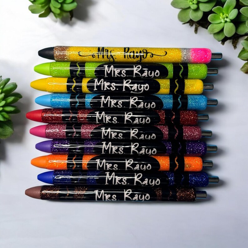 Puede incluir: Un conjunto de diez bol&iacute;grafos de purpurina de colores con etiquetas negras que dicen "Mrs. Rayo". Los bol&iacute;grafos est&aacute;n dispuestos en una pila, con el bol&iacute;grafo amarillo en la parte superior y el bol&iacute;grafo marr&oacute;n en la parte inferior.