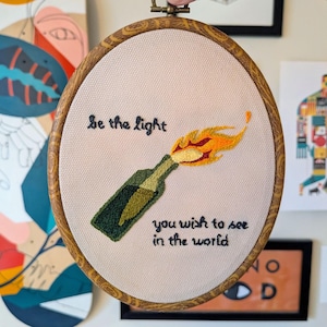 May include: Embroidered artwork in a wooden hoop. The design features a green bottle with a flame, and the text "be the light you wish to see in the world" in black thread. The background is a light beige fabric.
