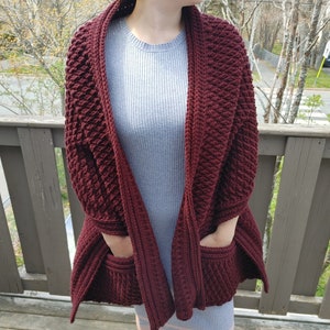 May include: A maroon crocheted cardigan with pockets. The cardigan is open in the front and has a textured stitch pattern.