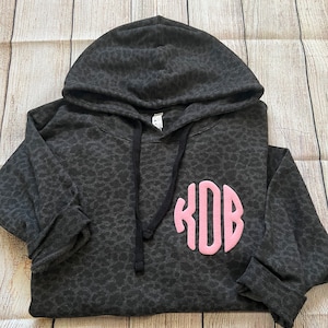 May include: Black hooded sweatshirt with a leopard print pattern and a pink monogram "KOB" on the front.