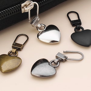 May include: Four heart-shaped zipper pulls in different finishes: silver, black, gold, and gunmetal. Each pull has a loop for attaching to a zipper.