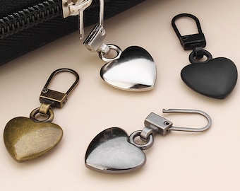 Könnte beinhalten: Vier herzförmige Reißverschlussanhänger in verschiedenen Ausführungen: Silber, Schwarz, Gold und Gunmetal. Jeder Anhänger hat eine Schlaufe zum Anbringen an einem Reißverschluss.