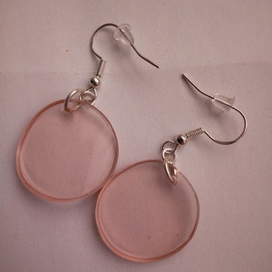 Pu&ograve; includere: Un paio di orecchini rotondi rosa con ganci d'argento. Gli orecchini sono realizzati in plastica trasparente con una tinta rosa.