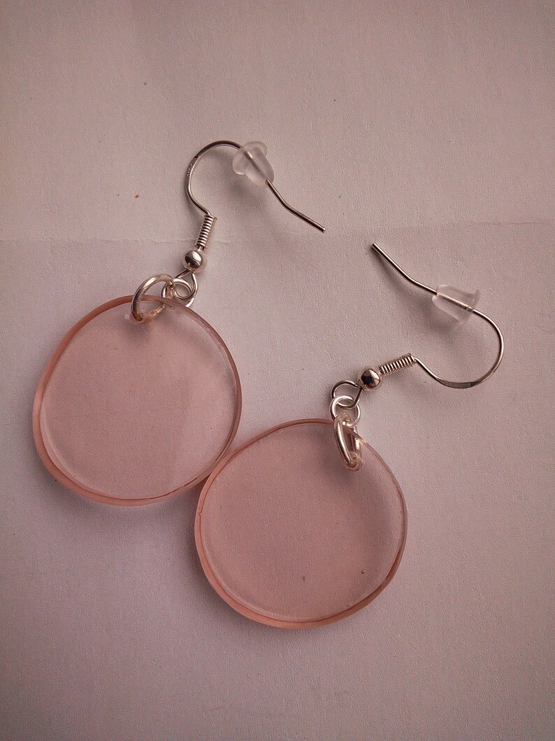 Pu&ograve; includere: Un paio di orecchini rotondi rosa con ganci d'argento. Gli orecchini sono realizzati in plastica trasparente con una tinta rosa.
