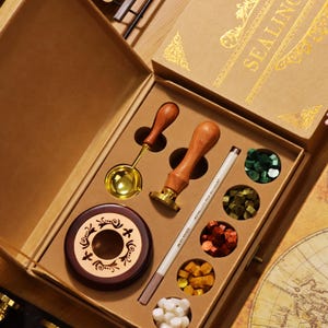 Puede incluir: Un kit de sellado de cera de estilo vintage en una caja marrón. El kit incluye un sello de cera con mango de madera, una cuchara para derretir, un bolígrafo metálico y cuentas de cera de colores. La caja está etiquetada "SEALING" en letras doradas.