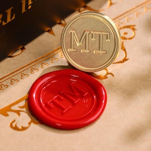 May include: A circular brass wax seal stamp featuring the engraved letters MT, positioned directly above a red wax seal impression that displays the embossed letters TM in a circular border, showcasing the stamp's design and its resulting mark.