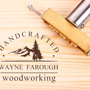 Może przedstawiać: Żelazko do znakowania wykonane z mosiądzu z napisem "HANDCRAFTED" i wzorem g&oacute;ry i sosny. Tekst "WAYNE FAROUGH WOODWORKING" jest r&oacute;wnież widoczny pod wzorem.