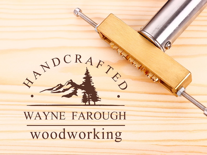 Może przedstawiać: Żelazko do znakowania wykonane z mosiądzu z napisem "HANDCRAFTED" i wzorem g&oacute;ry i sosny. Tekst "WAYNE FAROUGH WOODWORKING" jest r&oacute;wnież widoczny pod wzorem.