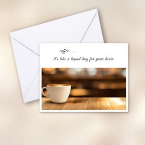May include: A greeting card with a white envelope. The card features a cup of coffee on a wooden table with the text "coffee... it's like a liquid hug for your brain". The coffee cup is white with a brown coffee and foam.