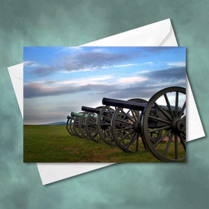 May include: A row of black cannons lined up on a grassy field with a blue sky and white clouds in the background.