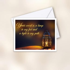 May include: A rectangular greeting card featuring a dark, glowing lantern with intricate metal cutouts and a warm light. The text reads: Your word is a lamp to my feet and a light to my path. Psalm 119:105. The card is placed on a white envelope.