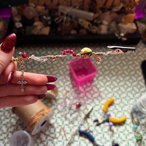 May include: A decorative alligator clip with gold wire, red beads, and a gold charm. A silver cross pendant hangs from the clip. The clip is embellished with a gold disc and clear and gold beads.