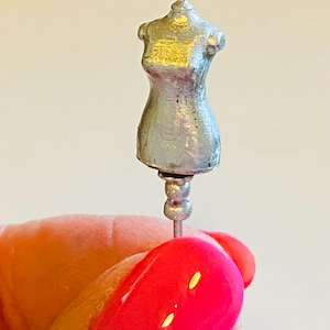 May include: A silver-colored miniature dress form with a round base and a pin on top. The dress form is held in a hand with pink nail polish.