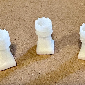 May include: Three white chess rook pieces are arranged on a brown surface. Each rook has a crenellated top and a square base. The rooks are made of a speckled, light-colored material.